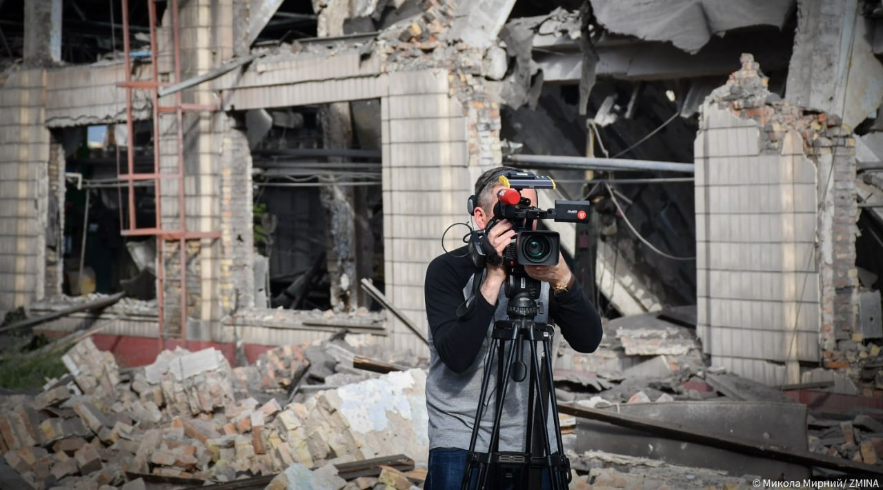 The operator stands with a video camera on a tripod in front of a ruined building. The ruins of the building, with damaged walls and broken bricks, are visible in the background.